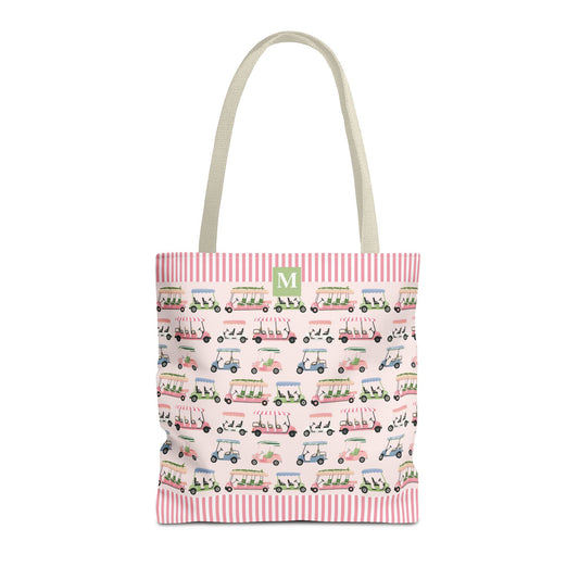 A canvas tote bag with a pink and white striped top border, featuring a repeating pattern of golf carts in pink, green, and blue on a pale pink background. The bag has light beige shoulder straps and a green square at the top with a white personalized initial.