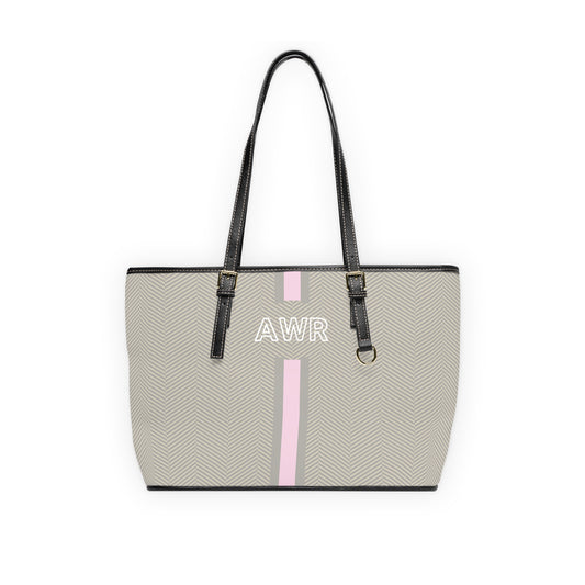A structured tote bag with a light neutral herringbone pattern, featuring a vertical pink stripe down the center, white monogram letters on the front, and black shoulder straps.